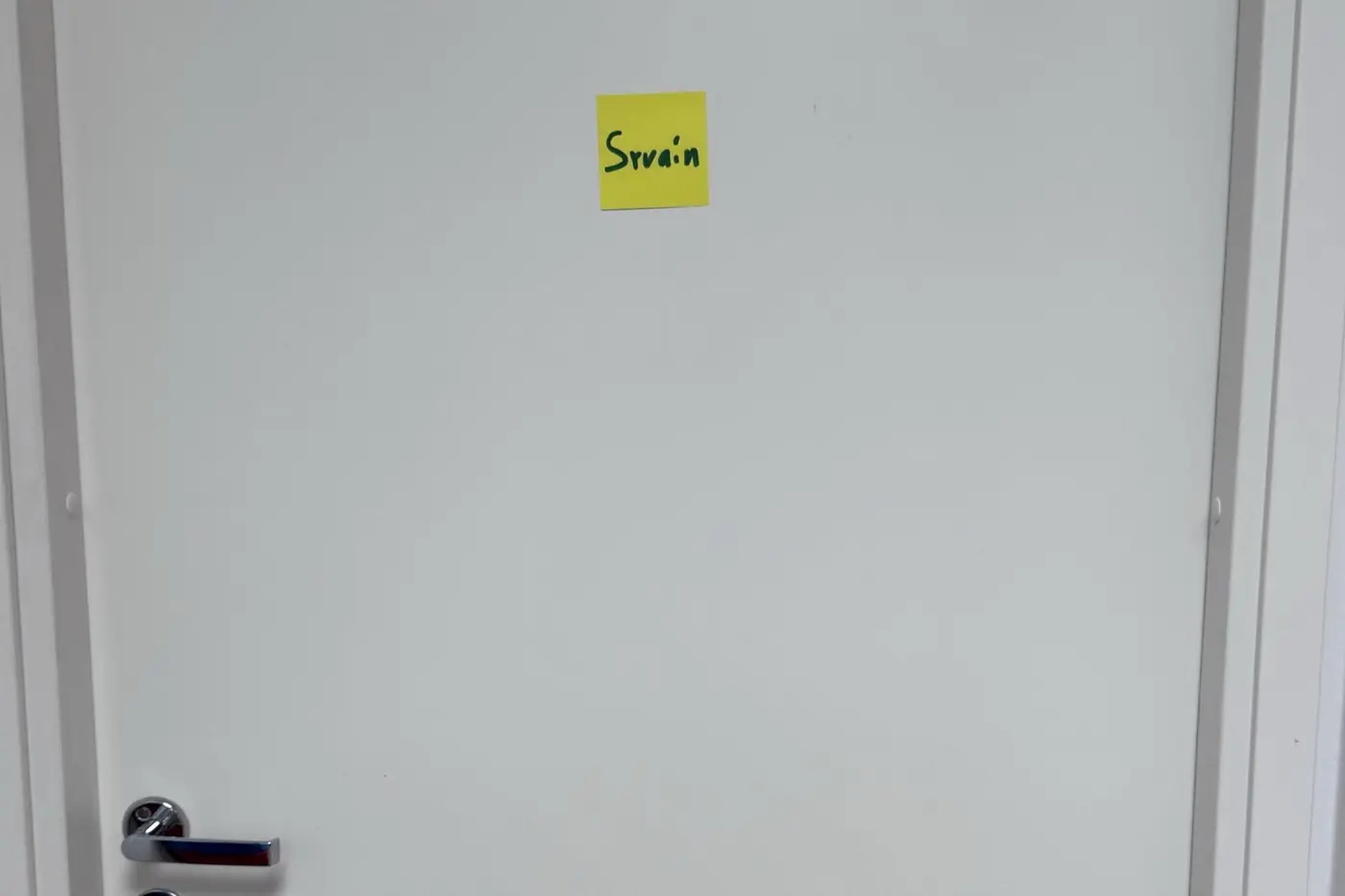 A plain white office door, empty except for a small yellow Post-it note hand-scrawled with our name.