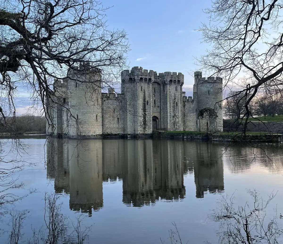 A medieval stone castle with round towers, encircled by a still moat that mirrors it.